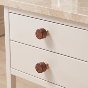 Pu&ograve; includere: Due pomelli cilindrici per mobili con frontali in legno marrone scuro e basi in metallo dorato. I pomelli sono fissati su un mobile bianco a due cassetti con un piano in marmo beige chiaro. La venatura del legno &egrave; visibile sulle superfici.