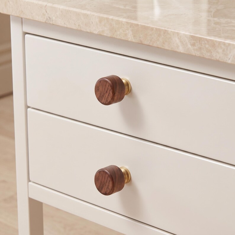 Pu&ograve; includere: Due pomelli cilindrici per mobili con frontali in legno marrone scuro e basi in metallo dorato. I pomelli sono fissati su un mobile bianco a due cassetti con un piano in marmo beige chiaro. La venatura del legno &egrave; visibile sulle superfici.
