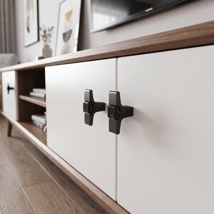Peut inclure: Un meuble TV bas compos&eacute; de portes d'armoire blanches et d'une structure en bois chaud. Les portes sont &eacute;quip&eacute;es de poign&eacute;es en m&eacute;tal noir en forme de T, dot&eacute;es d'une incrustation centrale en marbre noir et blanc vein&eacute;.