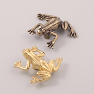 May include: Two frog-shaped cabinet knobs, one with a dark antique bronze finish and the other with a bright polished gold finish. Each knob features a detailed, realistic frog design with extended limbs and a central mounting post on the underside.