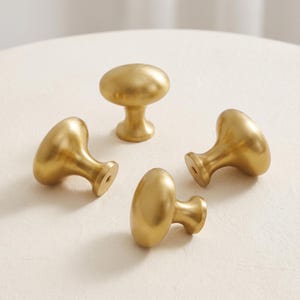 May include: A set of four gold-colored metal cabinet knobs with a smooth, matte finish. Each knob features a rounded, oval-shaped head and a tapered base with a visible screw hole at the bottom, designed for attachment to furniture or cabinetry.