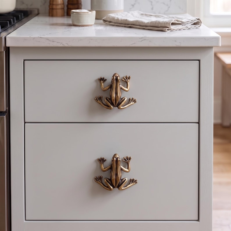 May include: Two antique brass-toned metal drawer knobs shaped like frogs, each featuring detailed limbs and a textured body. These decorative hardware pieces are mounted on the front of two stacked light gray kitchen cabinet drawers.