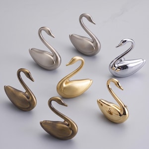 Puede incluir: Una colección de siete ganchos o pomos decorativos con forma de cisne, cada uno con un cuello y cuerpo curvados. El conjunto incluye varios acabados metálicos, como cromo pulido, oro cepillado, latón mate y tonos de bronce antiguo.