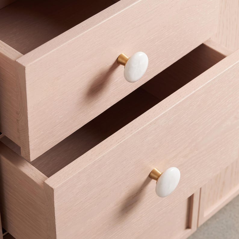 May include: A light pink wooden chest of drawers featuring two open drawers. Each drawer is fitted with a round, white knob attached to a gold-toned metal base. The wood grain is visible on the drawer fronts, showcasing a natural, light-toned finish.