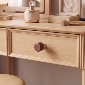 Pu&ograve; includere: Un tavolo da trucco in legno chiaro con un cassetto dotato di un pomello circolare in legno marrone scuro. Il frontale del cassetto presenta una modanatura rettangolare. La superficie ospita un flacone con tappo dorato, un portagioie e un pennello.