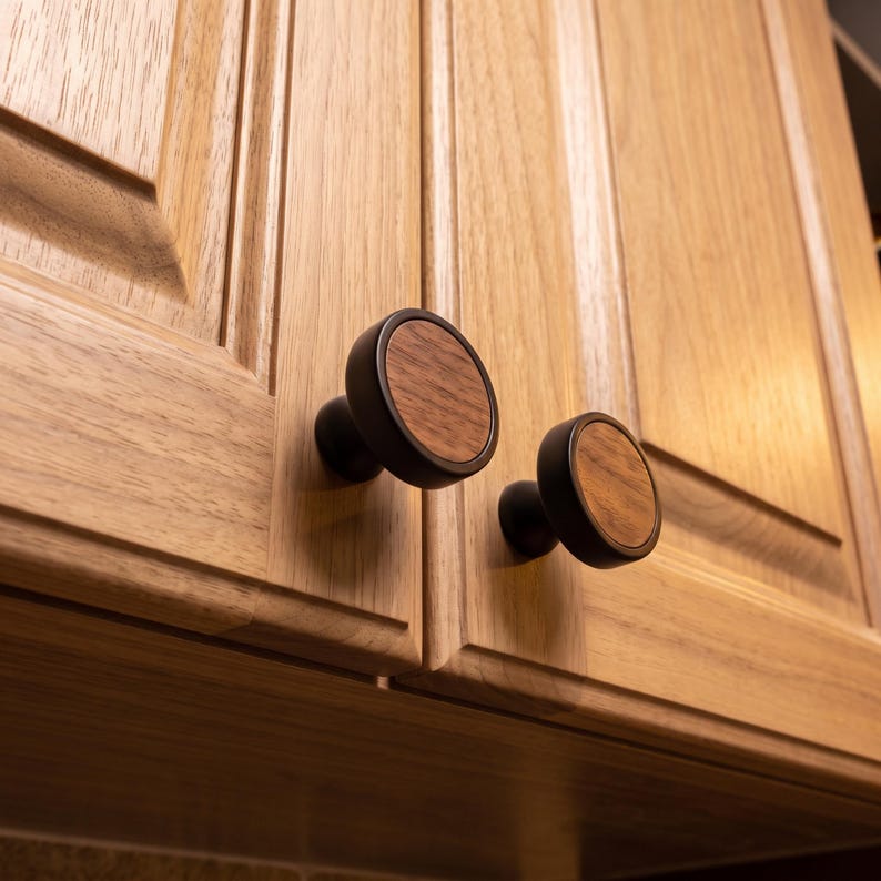 May include: Two circular cabinet knobs featuring a dark matte black metal frame surrounding a circular natural wood grain inlay. The knobs are mounted onto light brown wooden cabinet doors with vertical panel detailing and visible wood grain.