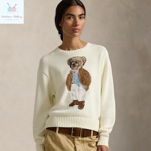 May include: Cream-colored sweater featuring an embroidered bear wearing glasses, a blazer, and trousers. The sweater has a ribbed hem and cuffs. A brown leather belt is visible. The brand logo is on the upper left corner.
