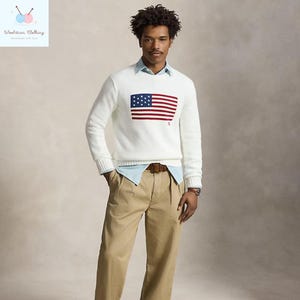 May include: A white V-neck sweater featuring a red, white, and blue American flag design on the chest. The sweater is paired with a light blue collared shirt, khaki pants, and a brown belt. The image also shows a logo in the top left corner.