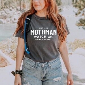 May include: A charcoal grey t-shirt featuring the white text "Mothman Watch Co. Point Pleasant". The shirt is paired with blue denim shorts. The image is set outdoors with a natural backdrop of rocks and water.