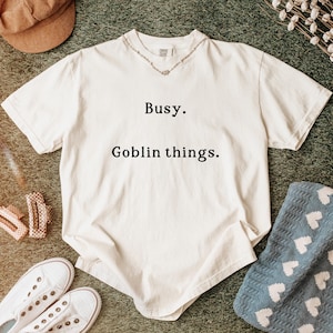 May include: Cream-colored t-shirt with the text "Busy. Goblin things." A brown corduroy cap, white sneakers, and a blue blanket with white hearts are also visible. A silver necklace is on the shirt.