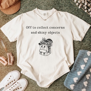 Puede incluir: Camiseta color crema con el texto "Off to collect concerns and shiny objects" sobre un gráfico de mochila llena de plantas y setas. La camiseta se combina con zapatillas blancas, pinzas para el pelo y una gorra marrón.