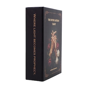 May include: A black tarot card box with gold lettering and floral accents. The box features the text "THE DIVINE ARTISAN TAROT" and "WHERE LIGHT BECOMES PROPHECY."