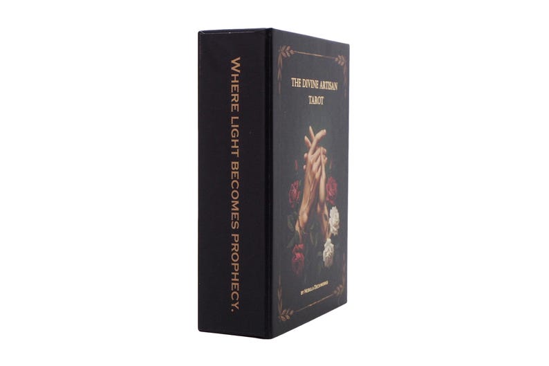 May include: A black tarot card box with gold lettering and floral accents. The box features the text "THE DIVINE ARTISAN TAROT" and "WHERE LIGHT BECOMES PROPHECY."
