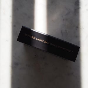 May include: A black rectangular box with gold lettering that reads "WHERE LIGHT BECOMES PROPHECY." The box is positioned diagonally on a marble surface, with shadows cast across the background.