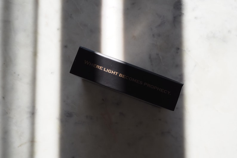 May include: A black rectangular box with gold lettering that reads "WHERE LIGHT BECOMES PROPHECY." The box is positioned diagonally on a marble surface, with shadows cast across the background.