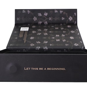 May include: A black box with an open lid, displaying a floral pattern on the inside. The box features the gold text "LET THIS BE A BEGINNING" on the front. The box is likely for gifting or storage of items.