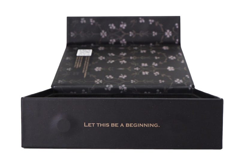 May include: A black box with an open lid, displaying a floral pattern on the inside. The box features the gold text "LET THIS BE A BEGINNING" on the front. The box is likely for gifting or storage of items.