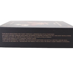 May include: A black tarot card deck box with text describing the deck's features. The text mentions luminous oil paintings, solitary figures, and serene gestures. The box is rectangular and sits on a white surface.
