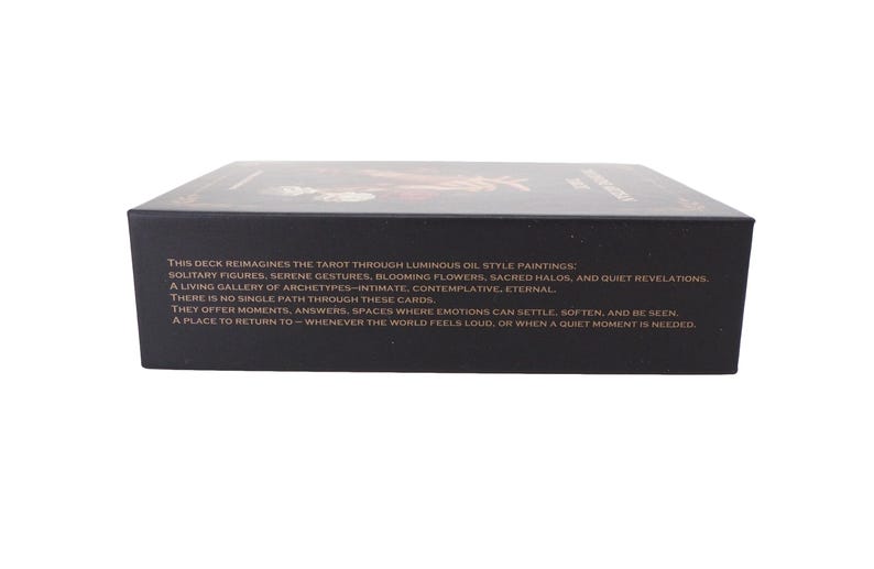May include: A black tarot card deck box with text describing the deck's features. The text mentions luminous oil paintings, solitary figures, and serene gestures. The box is rectangular and sits on a white surface.