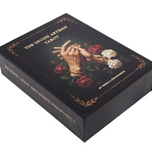 May include: A black tarot card box with gold lettering reading "THE DIVINE ARTISAN TAROT". The box features an illustration of two hands intertwined, surrounded by red and white roses. The text "WHERE LIGHT BECOMES PROPHECY" is also visible.