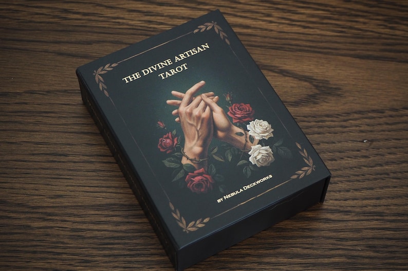 May include: A black tarot card box with the title "THE DIVINE ARTISAN TAROT" in gold lettering. The box features an illustration of clasped hands surrounded by red and white roses. The box is on a wooden surface.