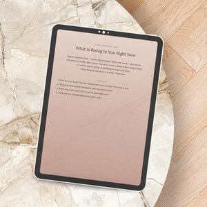 Peut inclure: Une tablette num&eacute;rique affichant le texte "What Is Rising In You Right Now" sur un fond rose clair. La tablette est pos&eacute;e sur une surface &agrave; motifs de marbre. Le texte comprend des invites &agrave; la r&eacute;flexion personnelle.