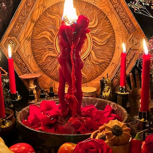 May include: A red candle shaped like two figures embracing, melting with a visible flame, set in a bowl filled with red rose petals. The scene includes surrounding red taper candles, fresh red roses, strawberries, and a carved wooden hexagonal plaque.
