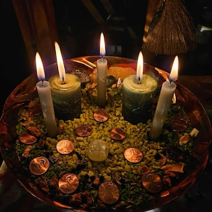 May include: A copper-colored bowl filled with small green stones, copper pennies, and a clear stone. Three thin grey candles and two thicker green candles are standing upright in the bowl, all with lit flames casting a warm glow on the contents.