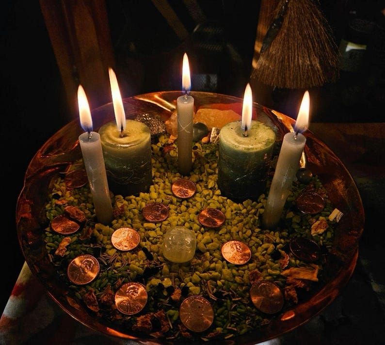 May include: A copper-colored bowl filled with small green stones, copper pennies, and a clear stone. Three thin grey candles and two thicker green candles are standing upright in the bowl, all with lit flames casting a warm glow on the contents.