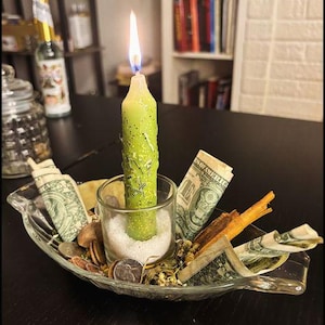 May include: A light green taper candle with gold glitter and embedded herbs burns inside a clear glass votive holder filled with white salt. The holder sits in a clear glass dish surrounded by rolled one-dollar bills, coins, and cinnamon sticks.
