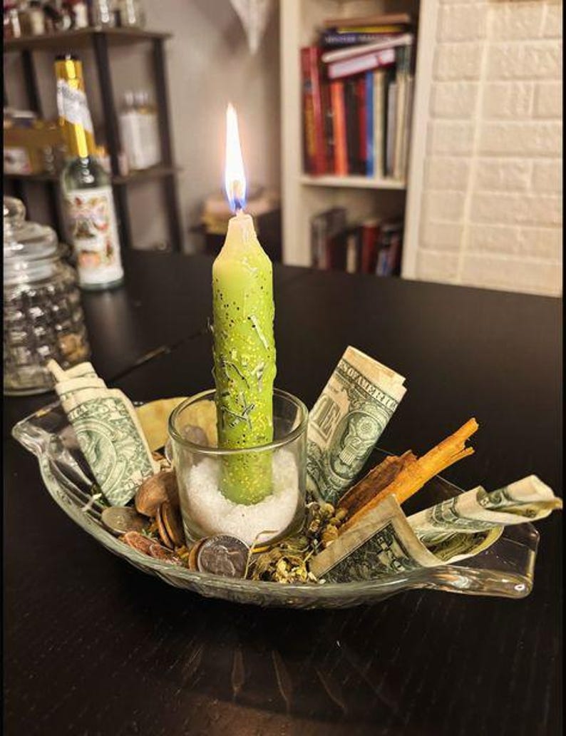 May include: A light green taper candle with gold glitter and embedded herbs burns inside a clear glass votive holder filled with white salt. The holder sits in a clear glass dish surrounded by rolled one-dollar bills, coins, and cinnamon sticks.