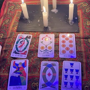 May include: A mystical scene featuring lit white candles and tarot cards arranged on a red patterned cloth. The cards display various illustrations and titles, including "EL MUNDO" and "LA PAPISA". The candles are positioned above the cards.