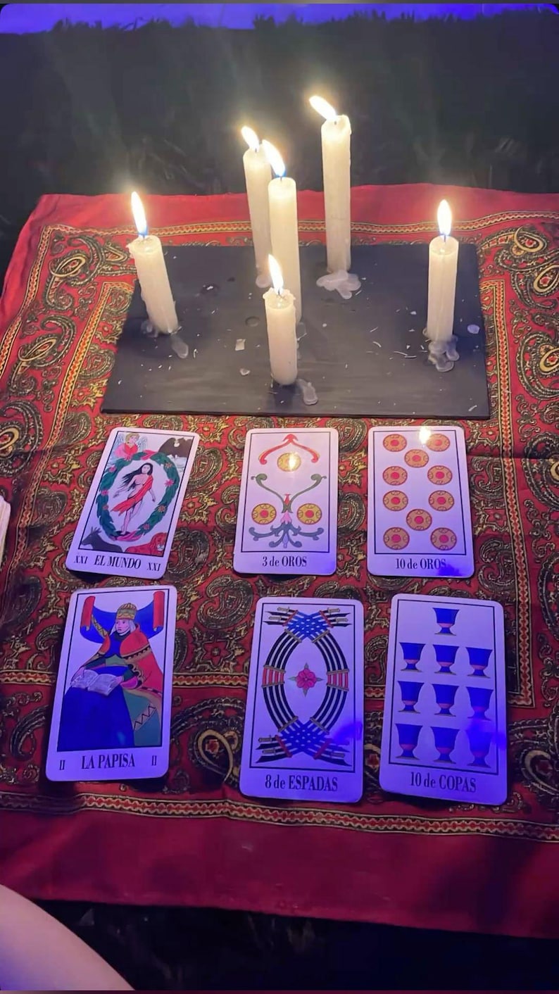 May include: A mystical scene featuring lit white candles and tarot cards arranged on a red patterned cloth. The cards display various illustrations and titles, including "EL MUNDO" and "LA PAPISA". The candles are positioned above the cards.
