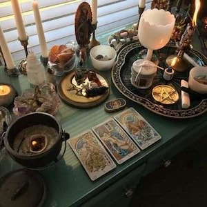 May include: A collection of spiritual items arranged on a table. The scene includes lit candles, tarot cards, a cauldron, a decorative tray, and various crystals. The items are arranged to create a mystical and esoteric atmosphere.