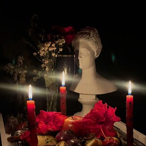 May include: A still life arrangement featuring a white bust, red candles, and a silver tray with fruit and flowers. The scene is dimly lit, with the flames of the candles providing the primary light source. The overall aesthetic is moody and artistic.