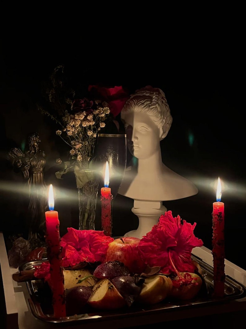 May include: A still life arrangement featuring a white bust, red candles, and a silver tray with fruit and flowers. The scene is dimly lit, with the flames of the candles providing the primary light source. The overall aesthetic is moody and artistic.