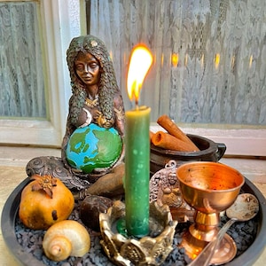 May include: A dark-toned figurine of a person with long hair cradling a blue and green globe, placed on a tray with black gravel. A green candle in a gold-toned holder, a copper chalice, a small spoon, a shell, a pomegranate, and cinnamon sticks.