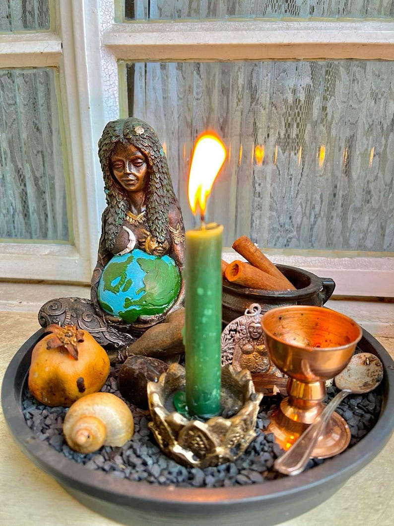 May include: A dark-toned figurine of a person with long hair cradling a blue and green globe, placed on a tray with black gravel. A green candle in a gold-toned holder, a copper chalice, a small spoon, a shell, a pomegranate, and cinnamon sticks.