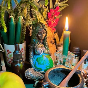 May include: A bronze-toned statue of a seated figure with hair made of leaves, cradling a globe painted with blue oceans and green continents. The figure is surrounded by green candles, a lit taper candle in a glass holder, a mortar and pestle, and a green apple.
