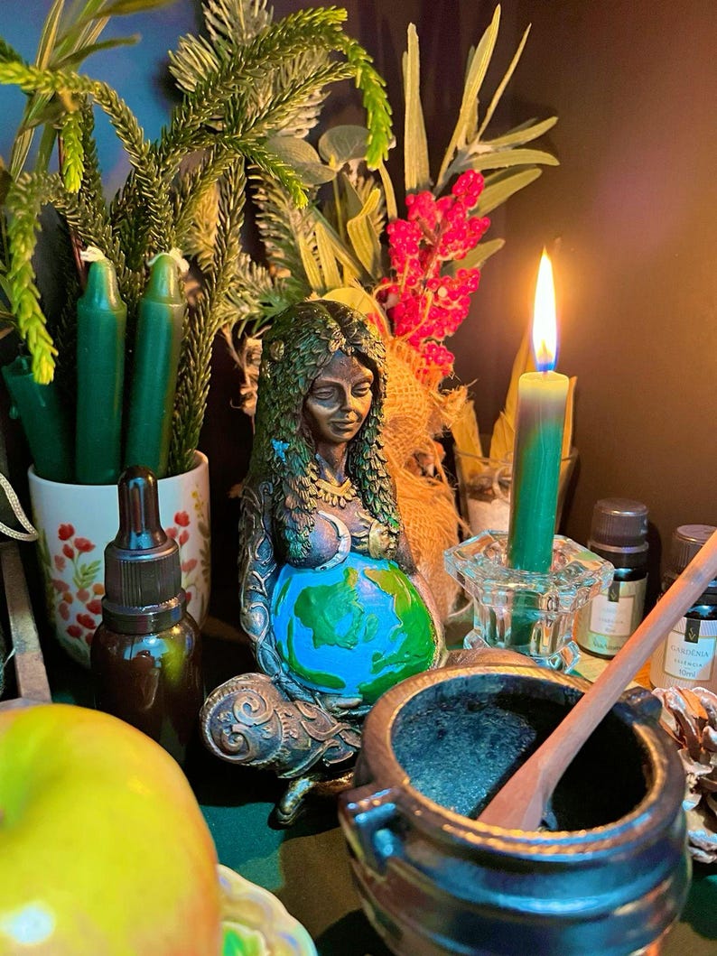 May include: A bronze-toned statue of a seated figure with hair made of leaves, cradling a globe painted with blue oceans and green continents. The figure is surrounded by green candles, a lit taper candle in a glass holder, a mortar and pestle, and a green apple.