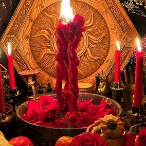 May include: A ritualistic scene featuring a large, red candle shaped like two figures, with flames atop. The candle is surrounded by red rose petals, candles, and a wooden, sun-shaped backdrop. Various fruits and flowers are also present.