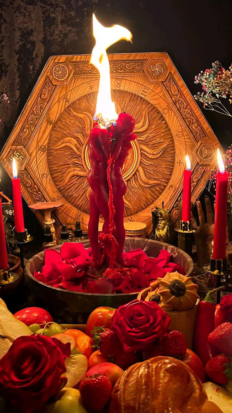 May include: A ritualistic scene featuring a large, red candle shaped like two figures, with flames atop. The candle is surrounded by red rose petals, candles, and a wooden, sun-shaped backdrop. Various fruits and flowers are also present.