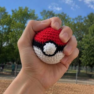 May include: A crocheted red and white ball with a black stripe and a white circle with a black outline. The ball is held in a hand against a background of trees and a blue sky.