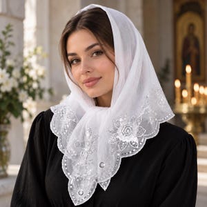 May include: A white head covering featuring intricate floral embroidery and delicate sequin detailing along the scalloped edges. The fabric is lightweight and semi-sheer, draped over the head and tied securely under the chin with a pointed front.