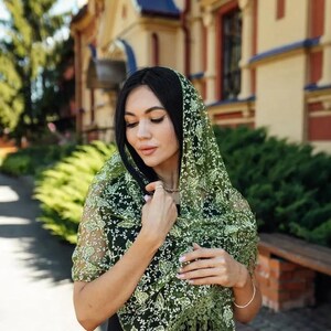 May include: A sheer green shawl with a delicate floral pattern and fringe trim. The shawl is draped over the shoulders, creating a soft, elegant look. The intricate design and color make it a versatile accessory.