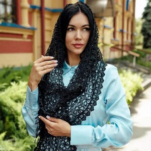 May include: A black lace shawl with a floral pattern is draped over a light blue collared shirt. The shawl is worn over the head and shoulders. The background features a building with yellow and red accents and green foliage.