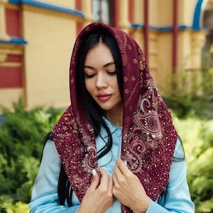 May include: A burgundy headscarf with an ornate paisley pattern, draped over a light blue blouse. The headscarf features a detailed design with floral and scroll elements. The image is taken outdoors.