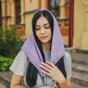 May include: A light purple headscarf featuring a delicate, tonal floral pattern. The square-shaped fabric is draped over the head, showcasing a soft texture and intricate, repeating botanical motifs across the entire surface of the lavender-colored accessory.