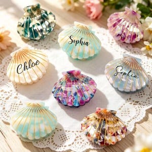 May include: A collection of eight shell-shaped hair clips made of colourful resin with gold-tone wire accents. Some clips feature custom names including Chloe and Sophia in black script lettering. The clips display various patterns like tortoiseshell, marble, and solid pastel colours.