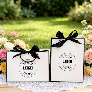 Può includere: Due sacchetti regalo bianchi con finiture nere e fiocchi neri. Ogni sacchetto presenta un'area logo circolare con la scritta "YOUR LOGO HERE". I sacchetti sono su una superficie di pizzo, circondati da fiori e verde.
